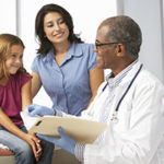 Smiling Doctor In Surgery Examining Young Girl With Mother.