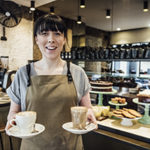 Female barista in her 20s serving coffee in cafe with cakes on display in background