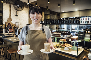 Female barista in her 20s serving coffee in cafe with cakes on display in background