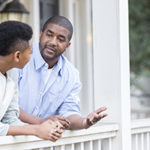 A father talks with his hands as he leans against the railing of his front porch with his preteen son and has a serious discussion.