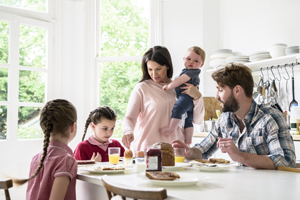 Two girls having breakfast with their dada as their mum carries their younger brother