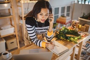 Working woman at gift shop. She wearing casual clothing, checking online shop with laptop and using mobile phone to receive new orders from customer