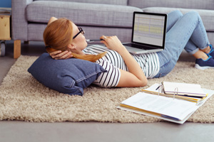 Young woman relaxing at home working or studying on a laptop computer lying on a rug on the floor with a notebook and binder alongside her, side view from behind