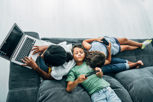 Mixed race family sitting on the sofa at home and using digital devices. African-american mother using laptop while her two children using smartphones.