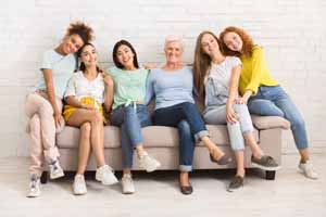Diverse Women Of Different Age Sitting On Sofa Smiling To Camera Indoor. Support Group Concept