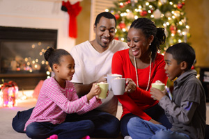 An African American family of four sharing hot cocoa on Christmas morning in the family living room next to the Christmas tree.
