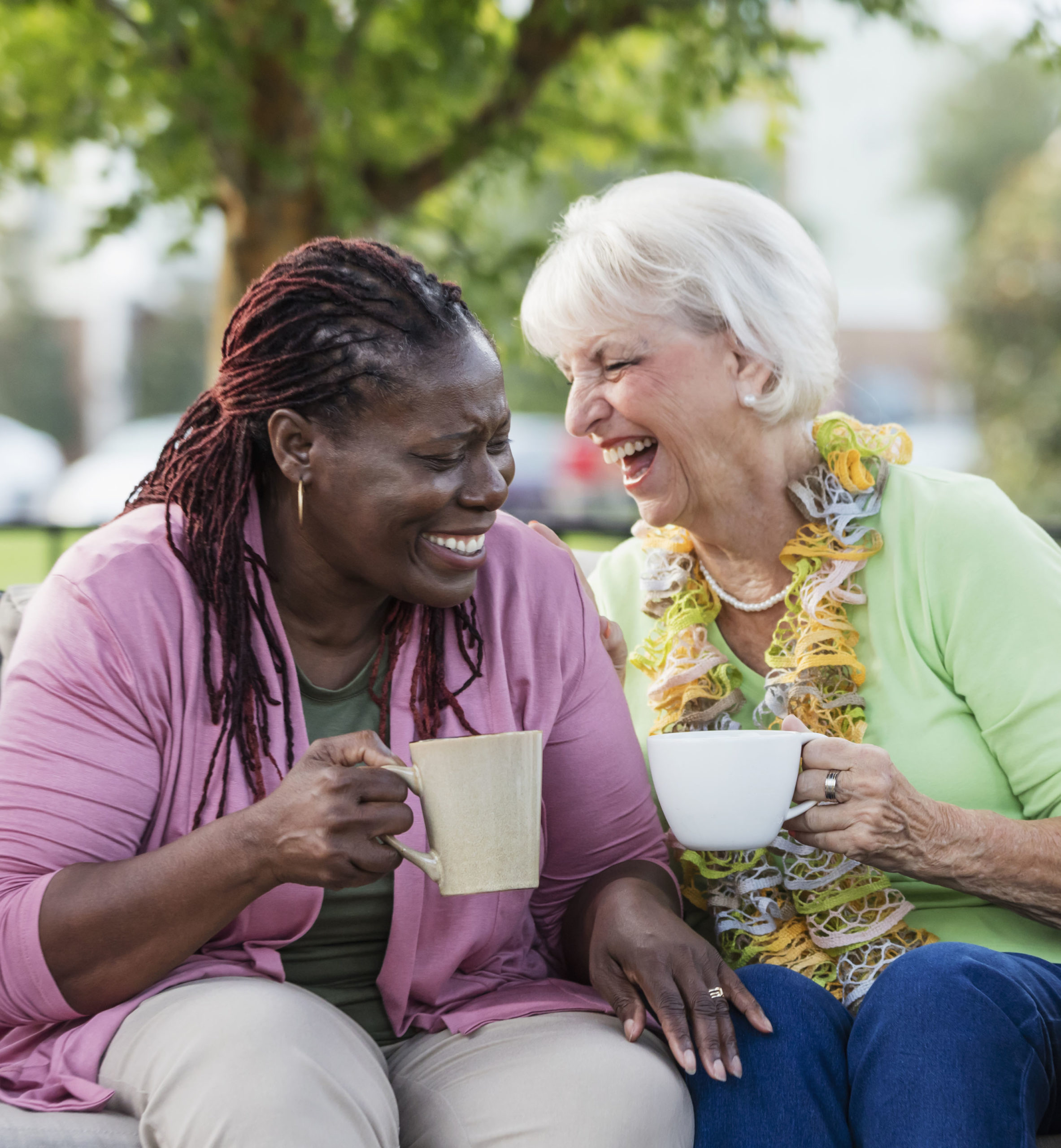 A senior woman in her 70s, drinking coffee and laughing with her African-American friend, a mature woman in her 50s. They are sitting outdoors on a patio sofa.