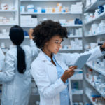 Shot of a young woman doing inventory in a pharmacy on a digital tablet with her colleague in the background