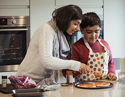 A mother is helping her autistic son spread the tomato sauce over the pizza dough in preparation for dinner.