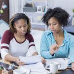 Young African American woman and mid adult African American woman work on a paper in a college dorm. They are using a digital tablet as well as a laptop. The bed is in the background.