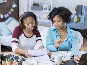 Young African American woman and mid adult African American woman work on a paper in a college dorm. They are using a digital tablet as well as a laptop. The bed is in the background.