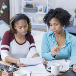 Young African American woman and mid adult African American woman work on a paper in a college dorm. They are using a digital tablet as well as a laptop. The bed is in the background.
