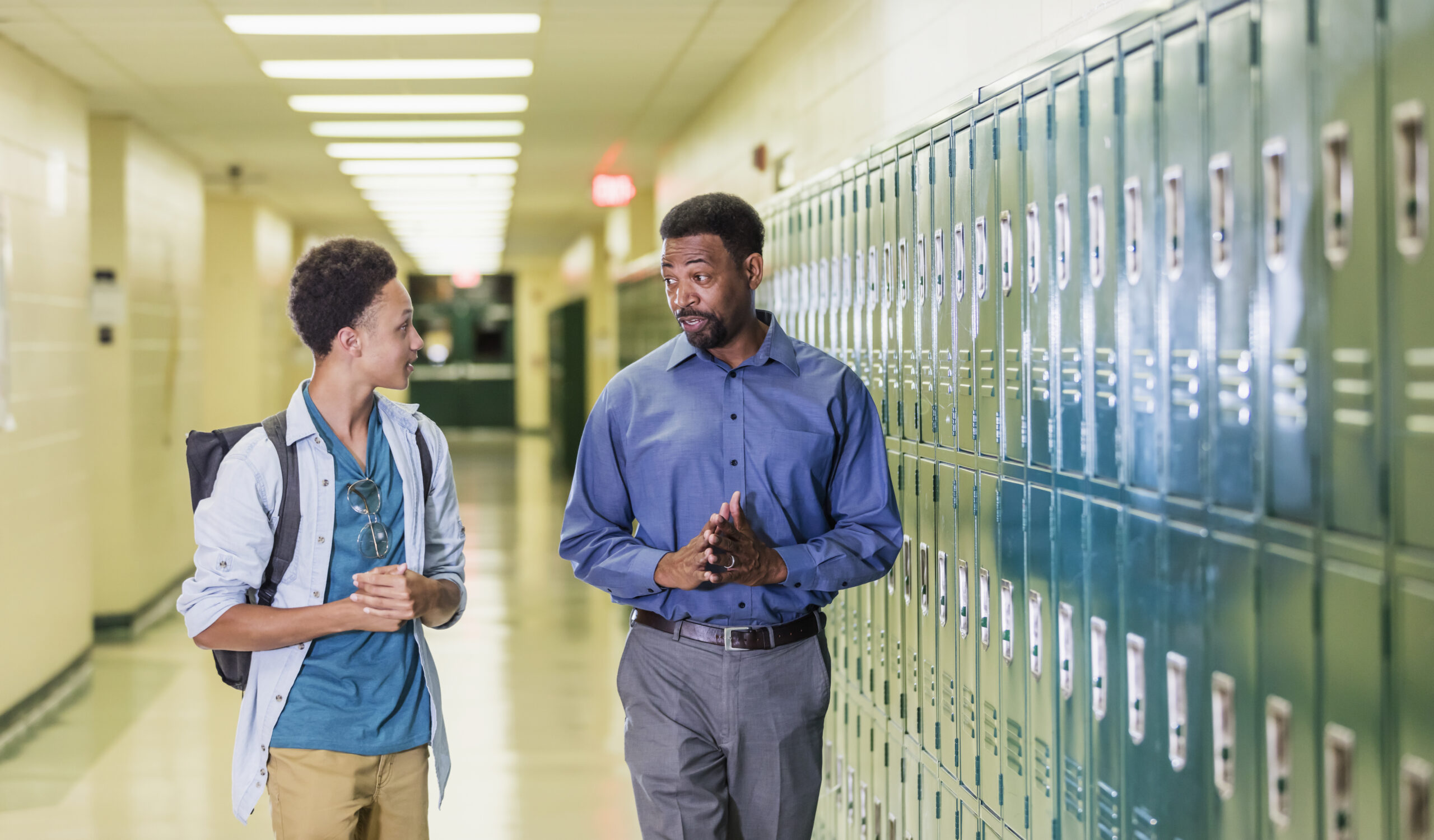 An African-American high school student walking with his teacher or school principal, in the hallway by a row of lockers. They are talking. The teacher has a serious expression on his face, a mature man in his 50s. The male student is an 18 year old teenage boy carrying a backpack.